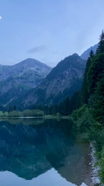 Video panorama during blue hour, overlooking a beautiful calm lake surrounded by steep mountains reflecting on the mirror-like water surface.
