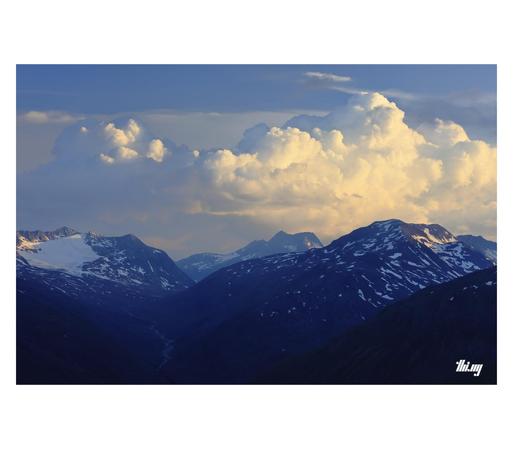 A high alpine glacial landscape with ice fields and snow patches during blue hour, a valley with its meandering glacial creek leading towards a pass in the distance.  Some of the summits are catching the last sun rays, as are the big cumulus clouds forming above.