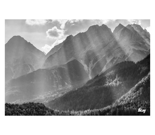B&W photo of an alpine landscape with dense forests in the valley and layers of tall, steep and rocky peaks higher up in the background. Intense light shafts are piercing the cloudy sky and hazy atmosphere, creating a beautiful light spectacle with some patches of the forest in direct sunlight.