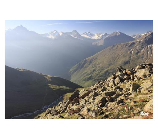 Photo of a high alpine landscape with three different valleys in bright hazy morning light. In the bottom left corner, still in the shade, a meandering creek is flowing down the rocky hanging valley formed by its former glacier. A small talus field in the sun, next to the path. Further in the distance much larger, deeper valleys, their floor occluded and only the upper sections illuminated. Glaciated peaks along the whole horizon, partially blanked out by intense bright light. Clear blue sky.