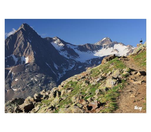 A small footpath, surrounded by talus fields, leading into the distance. Behind, on the other side of the invisible valley, some of the immense, imposing, glaciated peaks of the Kreuzkamm, specifically Talleitspitze (3406m) and Kreuzspitze (3455m). Not much is remaining of the formerly grand Eisferner in between these peaks. Clear blue sky.