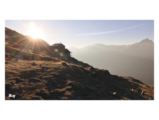 Wide-format photo of a mountain hut in a high alpine landscape, the sun rising just above the nearby ridge, creating a large sun star effect. Hazy atmosphere and some other mountains vaguely visible in the distance. Beautiful golden light. 