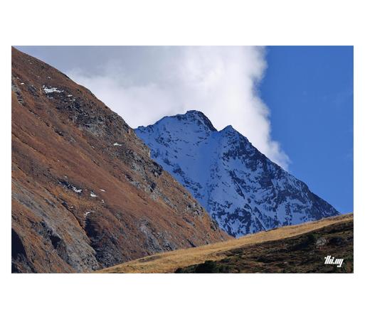 Abstract detail photo of three mountains and a huge cumulus cloud behind. Each mountain has a different color, slope and texture. The front one beige/brown grass. The mountain behind is steeper with grayish rocky cliffs and rusty brown grass. The mountain peak in the background is almost fully covered in snow, reflecting the blue sky.