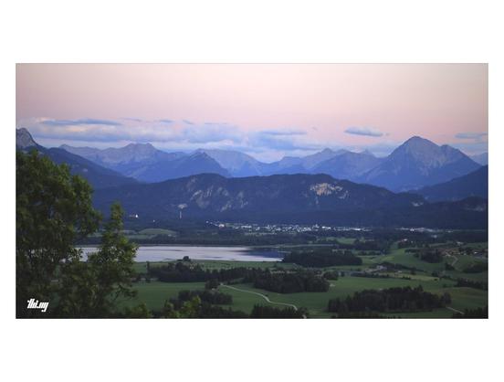 Wide-format view of the edge of the edge of the Alps near Füssen, with rolling hills, forests and a large lake in the foreground, the mountains of Tyrol in the back. Post sunset pink sky with blueish white clouds drifting above the mountain peaks how are also in blue mist/haze.