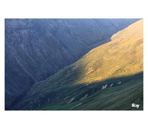 View down into a large, almost featureless alpine post-glacial valley without any vegetation other than grasses. The first sun of the day is illuminating some of the large slopes on the right hand side. The much steeper, almost vertical rocky cliffs on the left are still in the blueish shade, waterfalls running down every kilometer or so. The bottom of the valley is a deep and narrow canyon. The view is from around a 1000 meters above, covering an approx. 6km stretch of the valley. The canyon r…