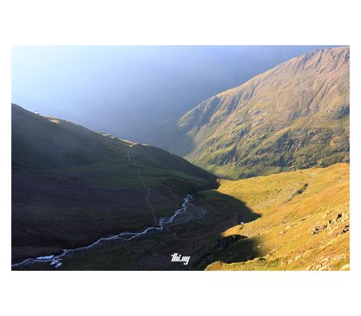 Bright super hazy morning light. A view down towards the confluence of two high alpine valleys with a third higher up one in the foreground, still 50% in shade, its glacier creek meandering through its rocky bed. A small hiking path crosses the creek and leads towards the drop off in the distance. 