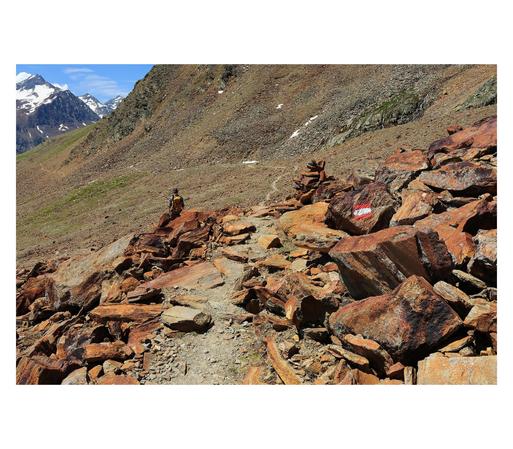 A high alpine path is leading through a scree field of intensely orange rusty rocks. Glaciated mountain peaks in the distance.