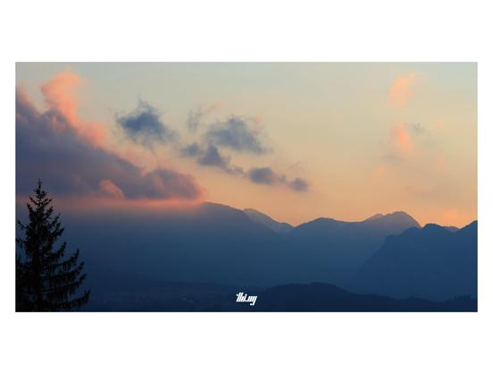 View of a mountain landscape post-sunset and onset of blue hour. A single cloud in an otherwise clear sky is illuminated in intense pinks, contrasting with the hazy silhouettes of the mountains below, all in shades of blue. The silhouette of a tree top in the bottom left corner.