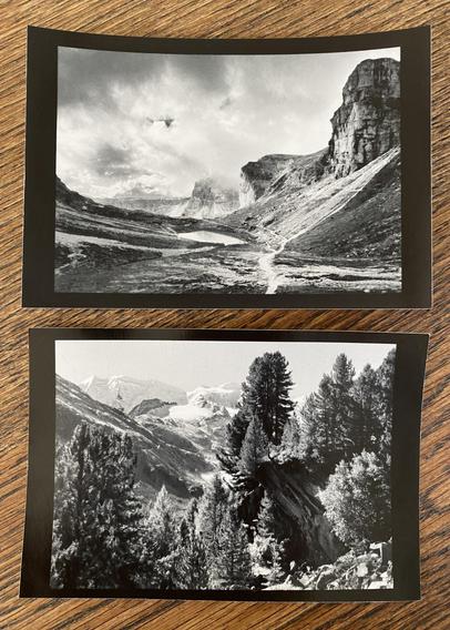 Phone capture of two small B&W prints with black border, depicting beautiful alpine mountain landscapes.

The top image shows a path in a high alpine valley leading into the distance past a small lake, surrounded by steep rock faces. Moody dark clouds forming.

The bottom image shows a large boulder within a group of swiss pines (also growing on top) in a talus field. Further in the distance the glaciated peaks of the Bernina range in bright sunshine.