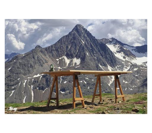 A wooden workbench standing close to the precipice in a high alpine landscape. A large mountain peak, still somewhat glaciated, is dominating the scene on the other side of the valley. Overcast moody sky, but the foreground is in the bright sun...