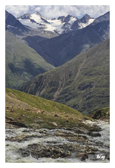 Portrait photo of a high alpine semi-glaciated landscape. The bottom third in the foreground shows a glacial creek rushing down through its stony bed into an increasingly steep valley/canyon (out of view), surrounded by eroded meadows growing on talus fields. Another ridge is running down sideways in mid distance. Further back another large valley with vast and steep grassy slopes and higher up a similar glacial creek where once still ice flowed, surrounded by its old moraines. Cloud shadows ar…