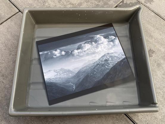 Phone picture of the same silver gelatin print, here still floating in a gray darkroom processing tray.