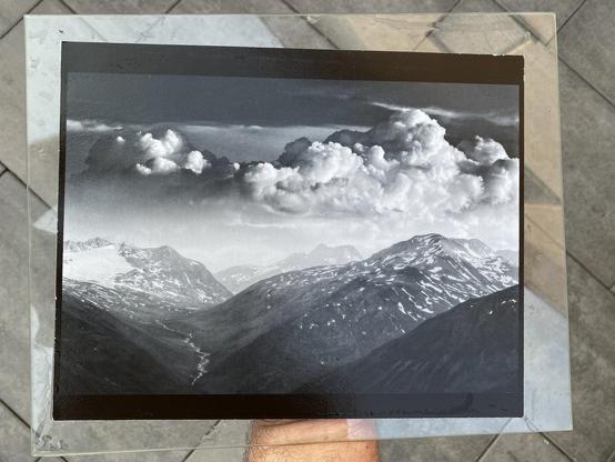 Phone picture of a person holding a still wet silver gelatin print on a glass sheet. The print shows a high alpine glacial mountain landscape with cumulus clouds floating above.