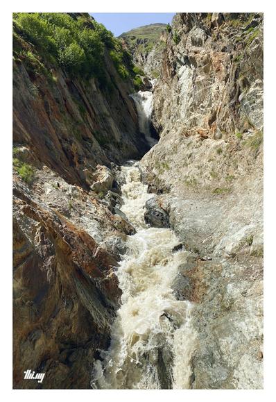 Portrait photo of a steep and narrow canyon carved by the outflow of a glacier higher up. The water is milky beige color, saturated with glacial flour. In the distance, higher up, is a mountain peak and a small section of the surrounding areas and meadows visible. Pale blue sky.