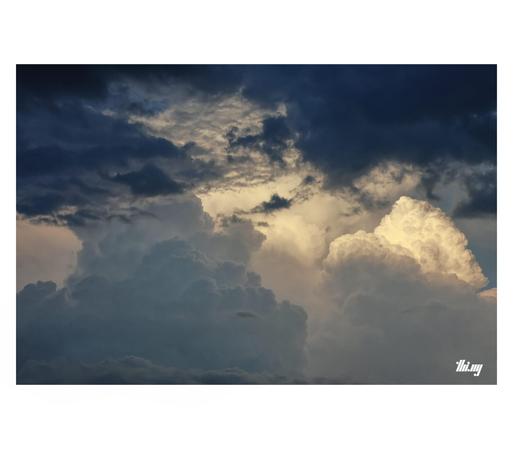 Detail photo of large sunlit cumulus clouds seen through a gap in the lower & dark cloud layer, which is also partially obscuring them. Beautiful soft peachy light on the main cloud. The bottom left cumulus has a subtle light bluish glow.