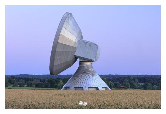 A large radio telescope (with a ~25 meters diameter parabola) seemingly standing in a corn field (it's actually behind). Layers of forest in the background, clear sky with a pink-blue gradient.