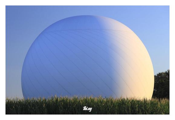 A large radio telescope covered with a ~50m diameter radome, illuminated by the warm morning sun and surrounded by a corn field. Some trees in the background, clear blue sky.
