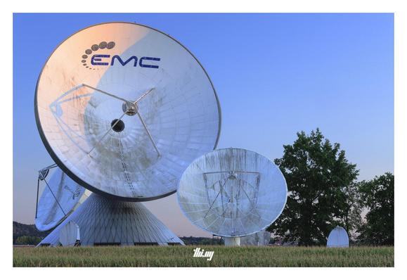 A group of radio telescopes of different sizes (from small to very large, ~25 meters diameter) seemingly standing next to a corn field and some trees. Clear sky with a pink-blue gradient. The rising sun is illuminating a part of the largest telescope's dish.