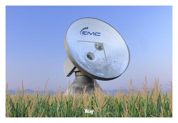 A large radio telescope (with a ~25 meters diameter parabola) seemingly standing in a corn field (it's actually behind). Some faint mountains in the background, clear sky with a pink-blue gradient.