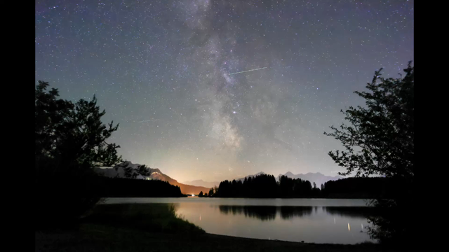 A 6 second time lapse of a view across a large lake with the night sky with the Milky Way and countless airplanes moving by. The scene is framed by the silhouettes of some bushes. The orange glow of the town of Füssen near the horizon. Faint mountain ridges further back.