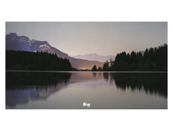 Wide-format long exposure photo of a large lake near the mountains in the background. The silhouette of a forest in mid distance. Behind it the orange glow of a small town (center) and the illuminated castle Neuschwanstein (left, castle itself not visible). Gray-bluish-purple starry sky. The water surface of the lake is smoothed by the long exposure, but the ripples caused by some wind near the forest is emphasizing the horizon line.