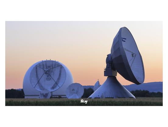 Wide-format view of some of the different antennas at Raisting Earth Station just before sunrise. Five storks are resting on the top edge of one of the parabolic dishes on the left, another single stork is sitting on another antenna further back. Clear pale peachy sky gradient. A small corn field in the foreground.