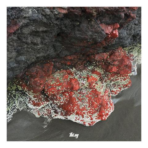 An exposed rock at the beach during low tide. Some parts of the rock are dark gray, others consisting of intensely red mineral with some white-greenish dotty overgrowth. Dark gray sands.
