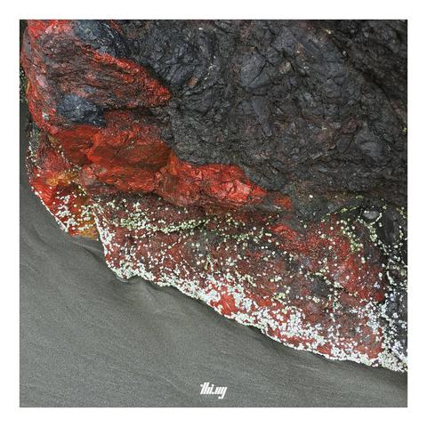 An exposed rock at the beach during low tide. Some parts of the rock are dark gray, others consisting of intensely red mineral with some white-greenish dotty overgrowth. Dark gray sands.