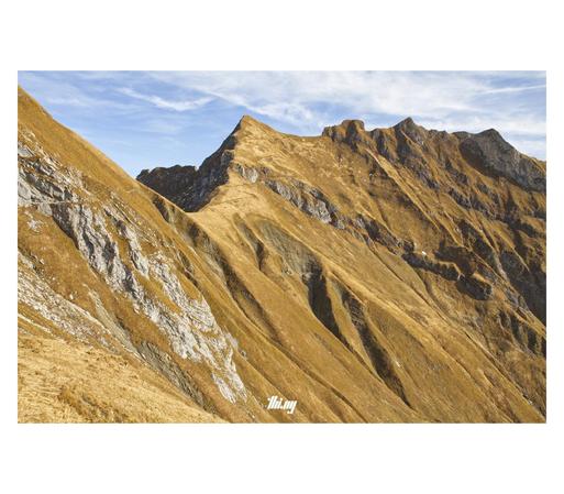 View of a mountain ridge with cliffs of light gray rocks and very steep grassy slopes dropping down towards the valley. The grass is in shades of golden brown. A small foot path is zigzagging up from a small pass towards the center peak. Pale blue sky with some cirrus clouds.