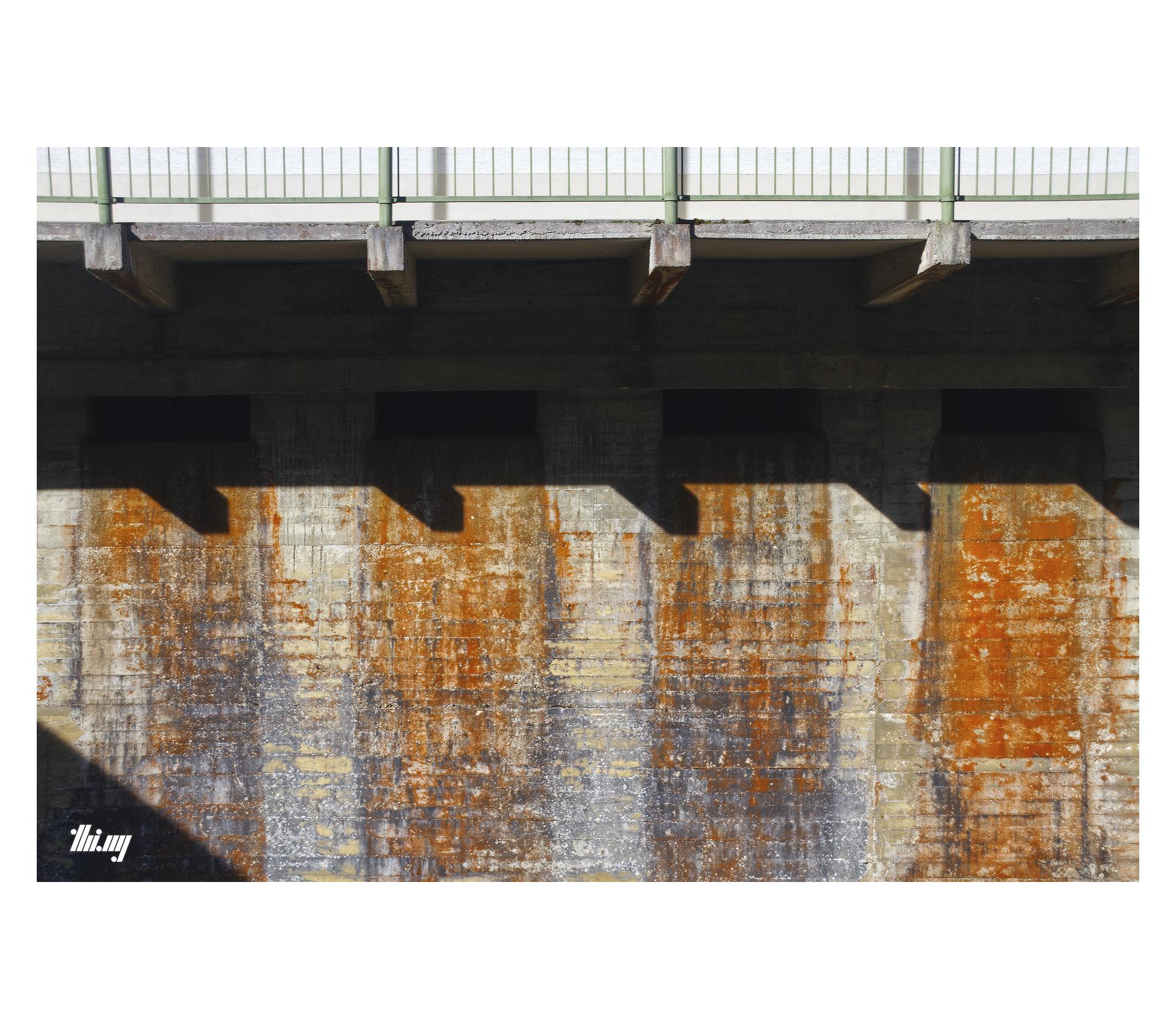 Photo of a cast concrete wall with four slots for emergency water outflow. The area below each slot is heavily coated in mostly orange lichen growth, together with other textures and shadows cast by other building structures (beams, railing), a highly abstract composition is created.