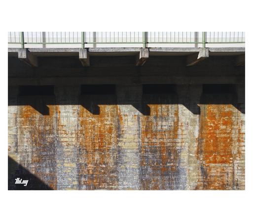 Photo of a cast concrete wall with four slots for emergency water outflow. The area below each slot is heavily coated in mostly orange lichen growth, together with other textures and shadows cast by other building structures (beams, railing), a highly abstract composition is created.