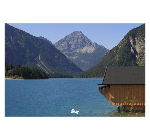 View of an intensely blue large lake surrounded by steep majestic mountains with large rock faces and dense forest further down. A small wooden boathouse on the right hand side. Sunny day with clear blue sky.