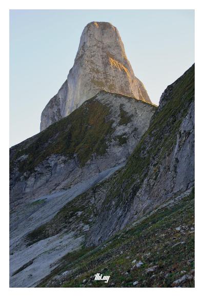 View of a rockneedle-like mountain peak (only looking like that from this side) in golden autumn afternoon light. In front, and more in the shade, another sharp ridgeline with colorful autumn grasses and talus fields. Clear bright sky.
