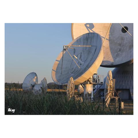 Photo of eight satellite dishes of varying sizes (from size of a person to 25 meters in diameter) standing near a corn field during sunrise. Parts of the dishes are illuminated in warm orange morning light. Clear blue sky.