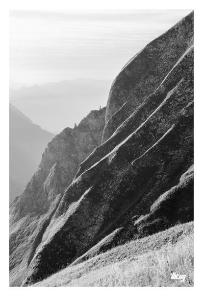 B&W portrait photo of steep, smoothly eroded rock slopes against a hazy background with faint layers of mountains. The rock face itself consists of several smooth layers of small ridges, leading up diagonally towards the top-right, their tops highlighted/emphasized by the high contrast early evening sun. Dried grasses in the foreground.