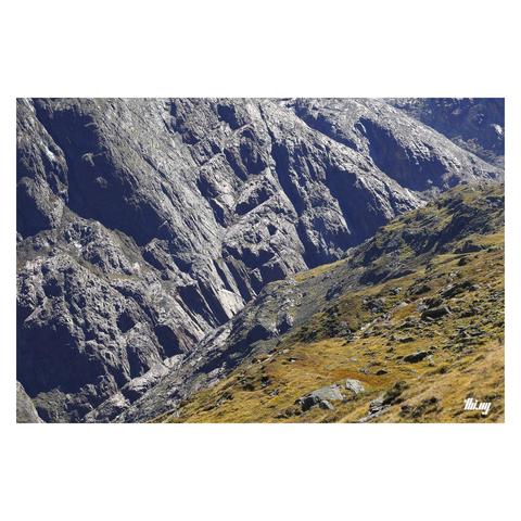 Photo of a deep canyon created by thousands of years of glacial abrasion with hundreds of meters tall cliffs of exposed bedrock (with cellular-automata-like patterns) on the opposite side and steep talus slopes, overgrown with grasses, on this side of the valley. It's early autumn and the vegetation in the foreground is already in shades of golden brown/orange, creating a color and textural contrast to the gigantic bare rock opposite.