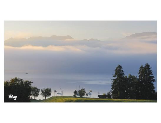 View of a large lake in the foothills of the Alps with a thick layer of low-hanging clouds illuminated by and glowing with the first soft peachy/pastel light of day. The clouds/fog are obscuring the horizon line and most of the mountains in the background. Only their top ridges are visible. Some meadows and trees surrounding a small pier with some boats in the foreground. A solitary fishing boat is in the middle of the lake, below the fog.