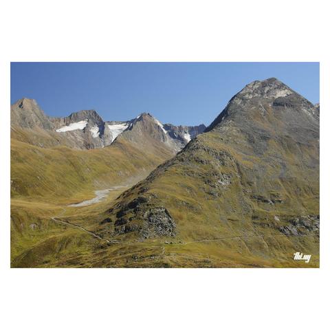 Photo of some smooth U-shaped glacial hanging side valleys in the central eastern Alps, parts of their remaining glacier fragments near the peaks the background. The lower slopes are covered in dry grasses in a mix of beautiful autumn colors. Talus fields and rocks of different colors higher up. Clear blue sky. A small gravel path is curving along the side of the main valley, passing two tiny-looking huts (near a talus field in the bottom center of the image).