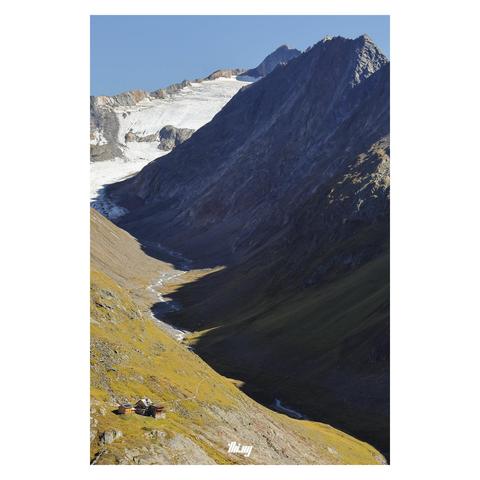 View of a mountain hut at the mouth of a high alpine glacial valley with steep rocky mountains on either side. The view shows the entire valley with the remaining glacier near the highest peak in the background and its outflow creek meandering through the valley. It's mid afternoon and the ridge on the right is casting harsh cold shadows. The hut and side buildings, on the other side of the valley, is still in the bright sun, surrounded by yellow/brown grasses among the bare scraped off rocks..…