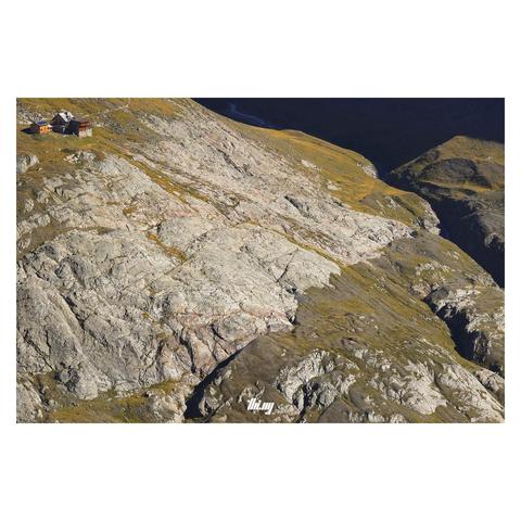 View of a mountain hut at the mouth and confluence of two high alpine glacial valleys with a large section of exposed bedrock dropping down toward a narrow canyon, carved by glacial creeks. It's late afternoon and the ridge on the right is casting harsh shadows. The hut and side buildings in the top left corner are still in the bright sun, surrounded by yellow/brown grasses among the bare scraped off rocks... No sky visible.