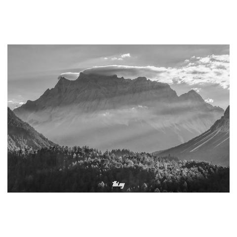 B&W photo of a large mountain massif with clouds getting stuck at its highest peaks. The view is framed by steep curved slopes of other mountains in mid distance. Strong diagonal light shafts piercing through the early morning haze, some of them also illuminating the tree tops of the dense forest in the center/foreground.