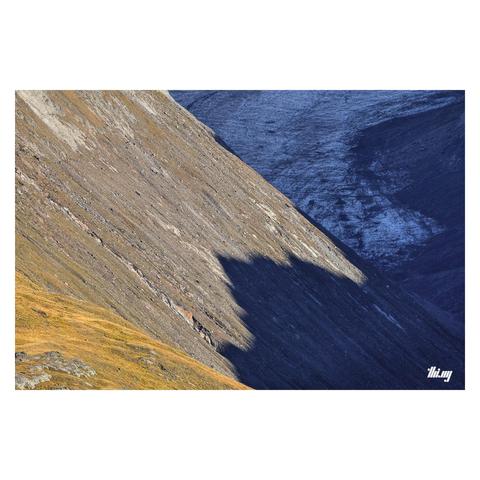 Cropped view deep into a glacial valley, with parts of the actual glacier visible in the back and steep sunlit talus slopes, partially covered in dried yellow grasses towards the foreground. The mountain peaks on the right side (out of view) are casting jagged hard shadows onto the slopes, further aiding the abstract character of this composition.