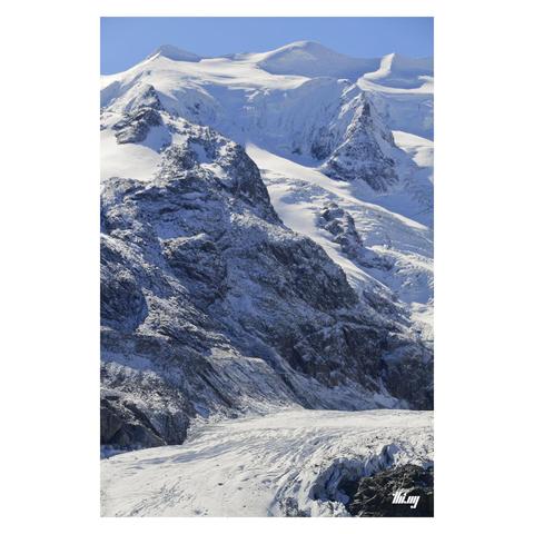 View of a large glacier flowing below its surrounding glaciated landscape of ~4000 meter peaks with huge ice falls and 1000+ meter tall rock faces dusted in fresh powder snow. Clear blue sky.