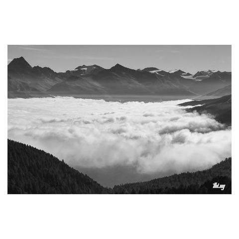 B&W photo of a large alpine valley covered by a thick layer of white clouds and thin strips of wispy mist above it. Majestic mountain peaks in the distance, some of them heavily glaciated. The view is from a mountain pass above the clouds. Steep slopes with dense forest in the foreground. 