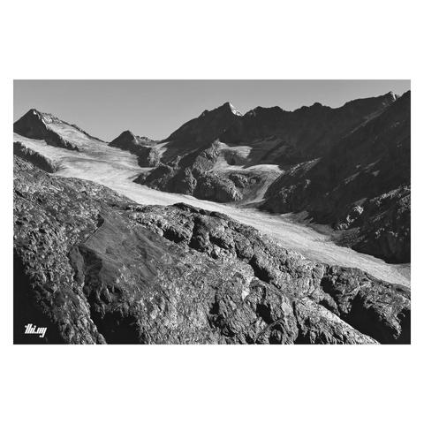B&W photo of a rocky high alpine landscape with a large glacier flowing diagonally across the image. A huge section/slope of exposed bed rock in the foreground, rocks glistening in the sun, leading down into a canyon (not shown). Further back, behind the ice flow, a ridge section of the Alpine divide with majestic peaks and several small side glaciers flowing into the main Ferner. Clear sky.