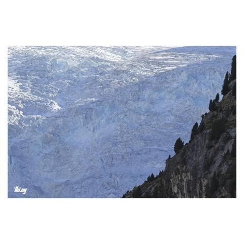 Frontal view of a section of a wall of almost a thousand meters of near vertical ice, a slowly shapeshifting maze of seracs, slowly flowing, fracturing, the light refracting and revealing the beautiful blue ice glow of the (dying) glacier... In the foreground the near silhouettes of a steep cliff section with some small Swiss pines.