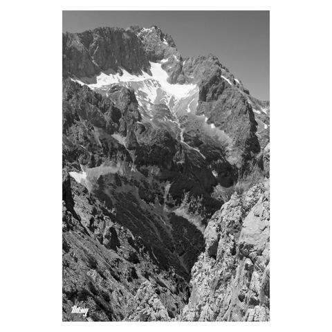 B&W portrait photo of a large alpine landscape with the Zugspitze peak (2962 m) and its glacier remnants below as central motif. The mountain drops off steeply (over the entire height of the image) into the valley below, talus fields giving way to fields of dwarf mountain pines and alpine meadows below. The valley bottom is not visible and obscured by some white limestone cliffs in the foreground, the view being from a very narrow pass between the rocks with steep drop offs directly ahead.