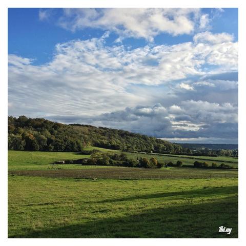 Photo of a typical English countryside with rolling hills, woodland ridges with trees in early autumn colors and big sky with layers of clouds (and some blue). The afternoon sun is casting long shadows in the foreground. A small hamlet in mid distance.