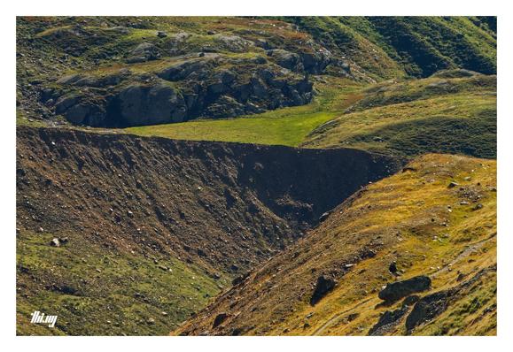 Abstract photograph of an alpine glacial valley. The cropped view omits any larger context and is purely focused on resulting shapes, colors and textures. The slopes are glacial deposits, partially overgrown with grasses in autumn colors (yellows, greens, browns). Some exposed bedrock and sharp-lined side moraine on the opposite side of the valley. A small path leading down into the valley in the bottom right corner.