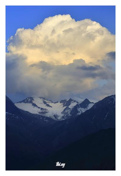 A single large cumulus cloud with golden yellow tint is floating above large glaciated mountain peaks and the already almost completely dark valley in front. The view is from high up and the valley stretches out below us in dark shades of blue.